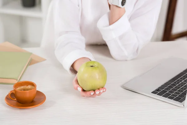 Abgeschnittene Ansicht einer Frau mit grünem Apfel in der Nähe von Laptop und Tasse im Büro — Stockfoto