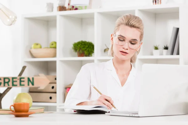 Femme écrivant dans un cahier près d'un ordinateur portable au bureau, concept d'économie de l'environnement — Photo de stock