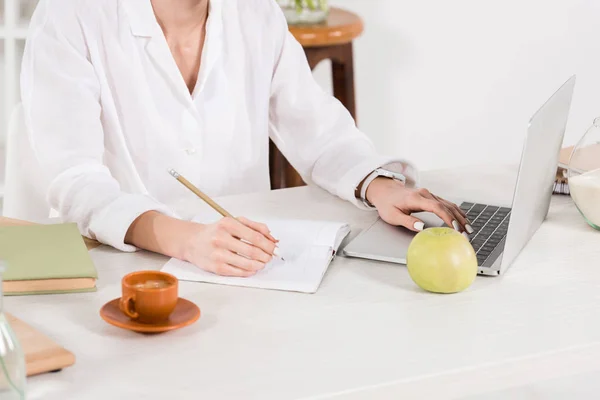 Ausgeschnittene Ansicht einer Frau, die in Notizbuch schreibt, während sie Laptop im Büro benutzt — Stockfoto
