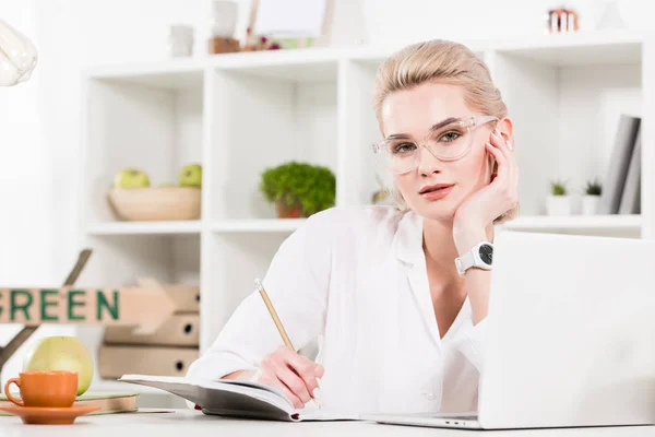 Woman in glasses writing in notebook near laptop in office, environmental saving concept — Stock Photo