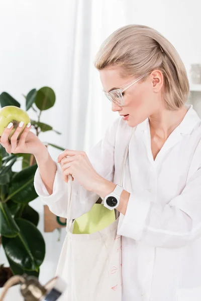 Beautiful businesswoman in glasses holding green apple and looking inside of eco bag, environmental saving concept — Stock Photo