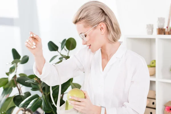 Cheerful businesswoman in glasses holding green apple and looking inside of eco bag, environmental saving concept — Stock Photo