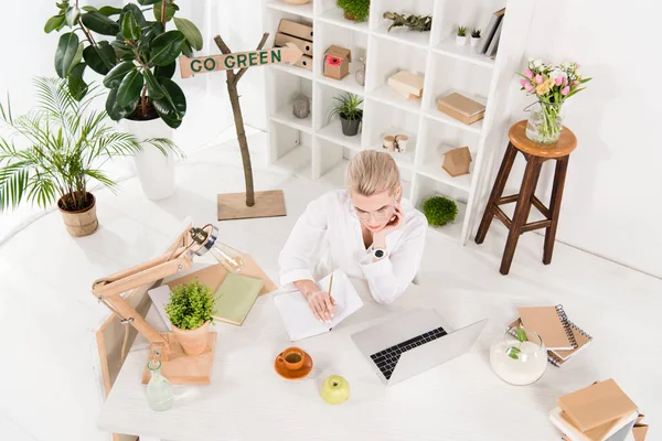 Вид сверху на предпринимательницу, пишущую в тетрадке рядом с ноутбуком с зеленым знаком сзади, концепция сохранения окружающей среды — стоковое фото