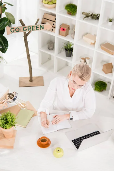 Overhead view of woman writing in notebook near laptop with go green sign behind, environmental saving concept — Stock Photo