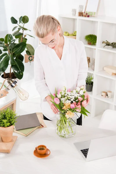 Веселая женщина смотрит на цветы в вазе возле ноутбука и чашки в офисе — стоковое фото