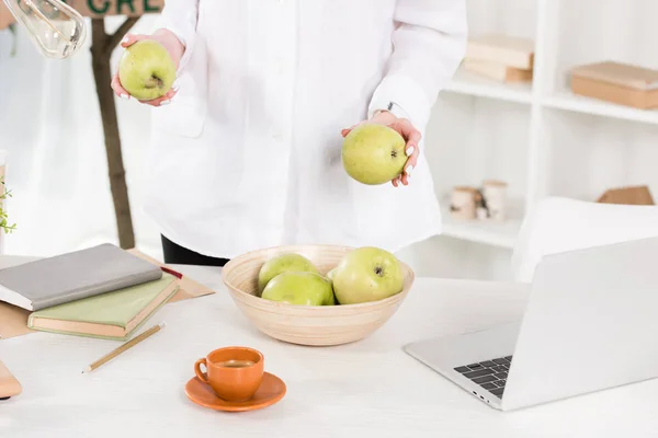 Обрізаний вид жінки, що тримає яблука в руках біля ноутбука в офісі — стокове фото