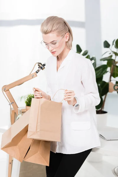 Hermosa mujer en gafas mirando las bolsas de compras en la oficina - foto de stock