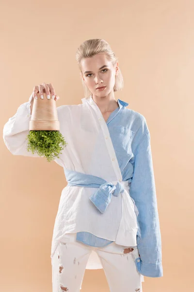 Femme tenant pot avec plante à l'envers tout en se tenant debout dans des vêtements écologiques isolés sur beige, concept d'économie de l'environnement — Photo de stock