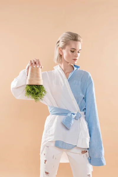Jovem mulher segurando panela com planta de cabeça para baixo, enquanto em pé em roupas ecológicas isolado no bege, conceito de poupança ambiental — Fotografia de Stock
