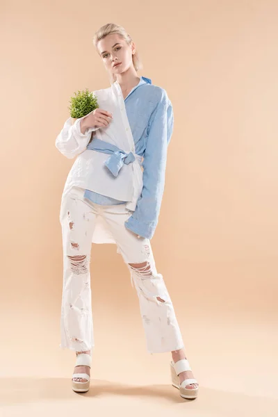 Hermosa mujer sosteniendo olla con planta y de pie en ropa ecológica aislada en beige, concepto de ahorro ambiental - foto de stock