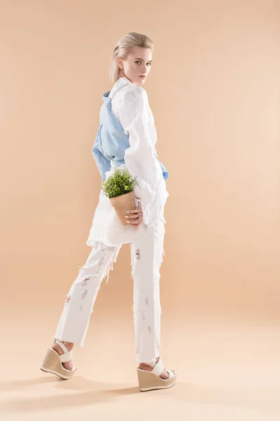 Красивая девушка держит горшок с растением и стоя в экологической одежде изолированы на бежевый, концепция сохранения окружающей среды — стоковое фото