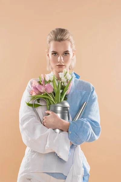 Femme embrassant arrosoir avec des fleurs et debout dans des vêtements écologiques isolés sur beige, concept d'économie de l'environnement — Photo de stock