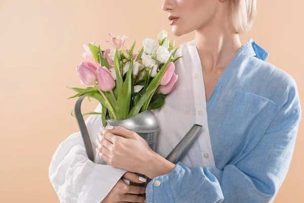 Abgeschnittene Ansicht einer jungen Frau, die Gießkanne mit Blumen in der Hand hält und in Öko-Kleidung isoliert auf beigem, umweltfreundlichem Konzept steht — Stockfoto