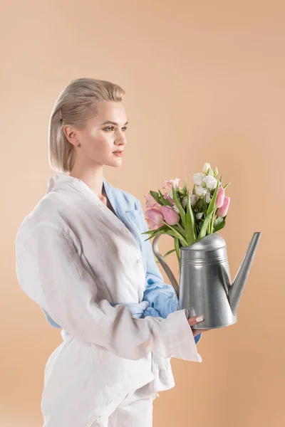Bela mulher segurando regador pode com flores e de pé em eco vestuário isolado no bege, conceito de poupança ambiental — Fotografia de Stock
