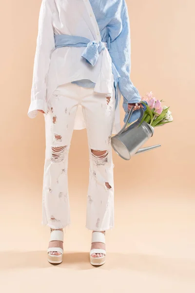 Обрезанный вид молодой женщины, держащей поливальную банку с цветами и стоящей в эко-одежде, изолированной на бежевом, экологически безопасной концепции — стоковое фото