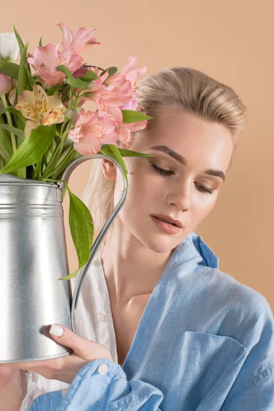 Atractiva mujer sosteniendo regadera con flores y de pie en la ropa ecológica aislado en beige, concepto de ahorro ambiental - foto de stock