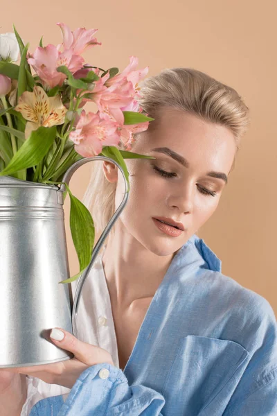 Attraktive Frau hält Gießkanne mit Blumen in der Hand und steht in Öko-Kleidung isoliert auf beigem, umweltfreundlichem Konzept — Stockfoto