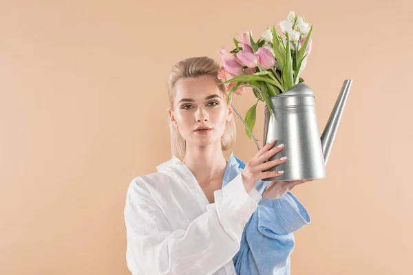 Hermosa chica sosteniendo regadera con flores y de pie en la ropa ecológica aislado en beige, concepto de ahorro ambiental - foto de stock