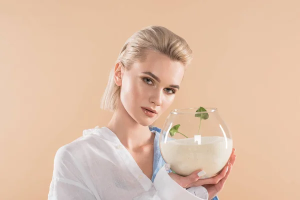 Beautiful girl holding fish bowl with sand and small green plant isolated on beige, environmental saving concept — Stock Photo
