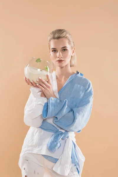 Hermosa mujer sosteniendo pecera con arena y pequeña planta verde, mientras que de pie en la ropa ecológica aislado en beige, concepto de ahorro ambiental - foto de stock