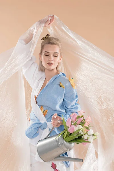 Mujer sosteniendo polietileno y mirando regadera con flores mientras está de pie en la ropa ecológica aislado en beige, concepto de ahorro ambiental - foto de stock