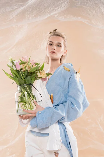 Menina atraente de pé com borboletas em roupas ecológicas e segurando vaso de vidro com flores em fundo bege, conceito de poupança ambiental — Fotografia de Stock