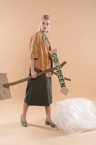 Blonde girl wearing eco clothing standing near polyethylene and holding wooden sign with go green lettering on beige background, environmental saving concept — Stock Photo