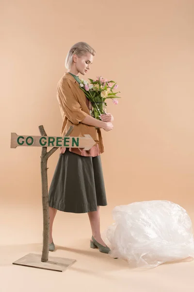 Rubia chica en eco ropa sosteniendo jarrón con flores cerca de signo con ir letras verdes sobre fondo beige, concepto de ahorro ambiental - foto de stock