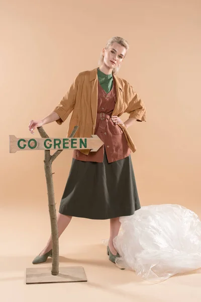 Attraente ragazza in piedi in eco abbigliamento vicino polietilene e andare segno verde su sfondo beige, concetto di risparmio ambientale — Foto stock