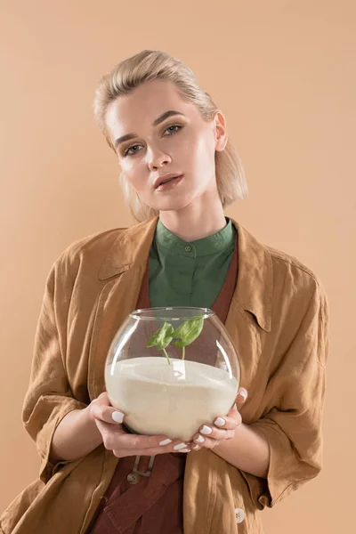 Hermosa mujer rubia sosteniendo pecera con arena y pequeña planta verde, mientras que de pie en la ropa ecológica aislado en beige, concepto de ahorro ambiental - foto de stock