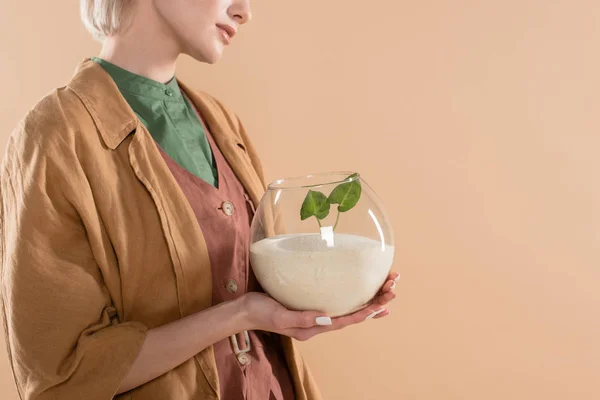 Vista recortada de la mujer sosteniendo pecera con arena y pequeña planta verde mientras está de pie en la ropa ecológica aislado en beige, concepto de ahorro ambiental - foto de stock