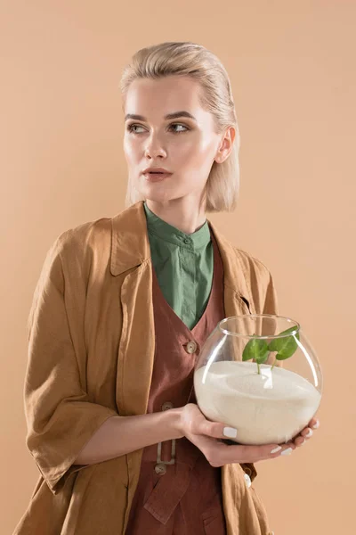 Mujer rubia sosteniendo pecera con arena y pequeña planta verde mientras está de pie en ropa ecológica aislada en beige, concepto de ahorro ambiental - foto de stock