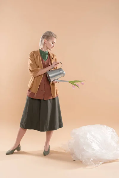 Blondes Mädchen in Öko-Kleidung hält Gießkanne mit Blumen in der Nähe von Polyethylen auf beigem Hintergrund, Umweltschutzkonzept — Stockfoto