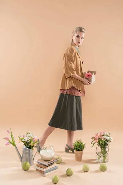 Atractiva mujer sosteniendo casa de cartón con flores cerca de plantas y flores sobre fondo beige, concepto de ahorro ambiental - foto de stock