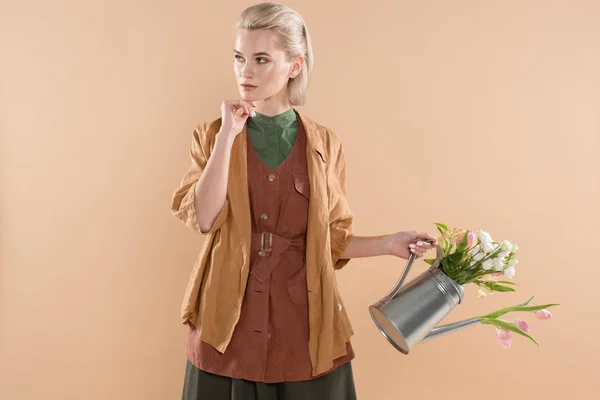 Chica pensativa en ropa ecológica sosteniendo regadera con flores aisladas sobre fondo beige, concepto de ahorro ambiental - foto de stock