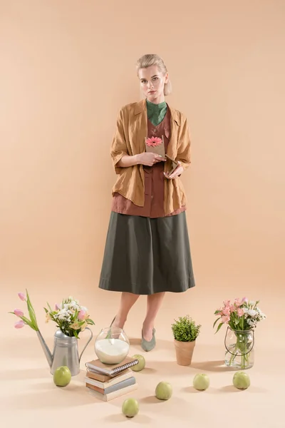 Donna bionda che tiene casa di cartone con fiore vicino a piante e fiori su sfondo beige, concetto di risparmio ambientale — Foto stock