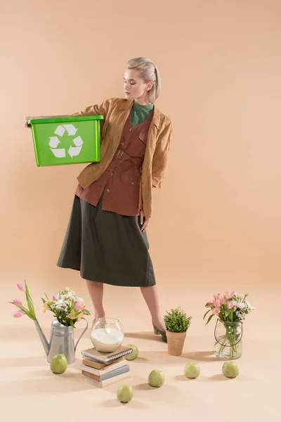 Mujer rubia sosteniendo caja de reciclaje cerca de plantas y flores sobre fondo beige, concepto de ahorro ambiental - foto de stock