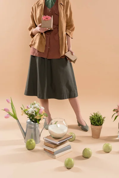 Обрезанный вид женщины, держащей картонный домик с цветами рядом с растениями и цветами на бежевом фоне, концепция охраны окружающей среды — стоковое фото