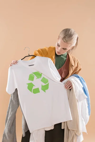 Mujer joven mirando la ropa ecológica con el cartel de reciclaje en la percha aislada en beige, concepto de ahorro ambiental - foto de stock