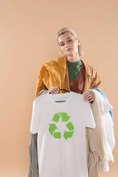 Hermosa mujer joven sosteniendo ropa ecológica con cartel de reciclaje en la percha aislada en beige, concepto de ahorro ambiental - foto de stock