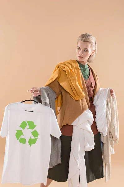 Jovem segurando roupas ecológicas com sinal de reciclagem no cabide isolado no bege, conceito de economia ambiental — Fotografia de Stock