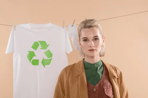 Selective focus of blonde woman standing near socks and t-shirt with recycling sign on clothesline isolated on beige, environmental saving concept — Stock Photo