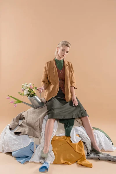 Chica rubia sentada en la pila de ropa y sosteniendo regadera con flores sobre fondo beige, concepto de ahorro ambiental - foto de stock