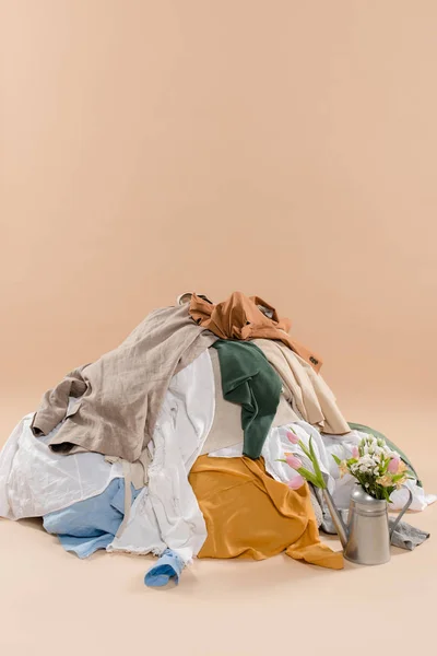 Стопка одежды возле лейки с цветами на бежевом фоне, концепция сохранения окружающей среды — стоковое фото