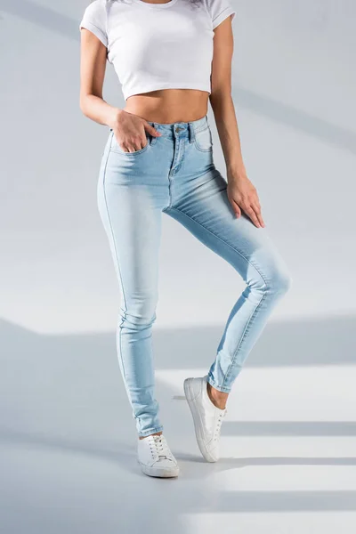 Обрезанный вид сексуальной девушки в синих джинсах, стоящей с рукой в кармане на сером фоне — стоковое фото