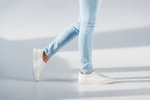 Partial view of woman in blue jeans and white shoes on grey background — Stock Photo