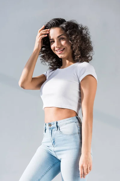 Chica alegre en jeans tocando el pelo rizado sobre fondo gris - foto de stock