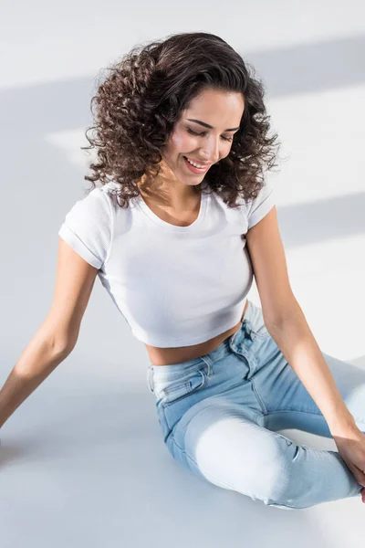 Bella ragazza riccia in jeans seduta sul pavimento con il sorriso — Foto stock