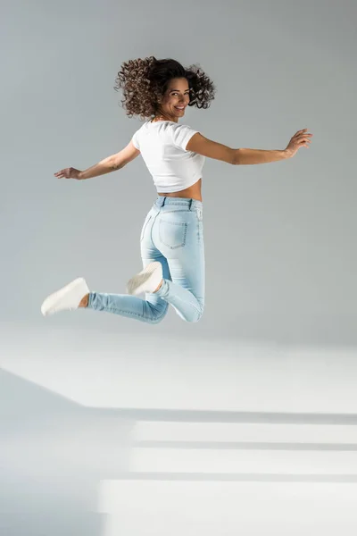 Femme bouclée riante en jeans sautant sur fond gris — Photo de stock