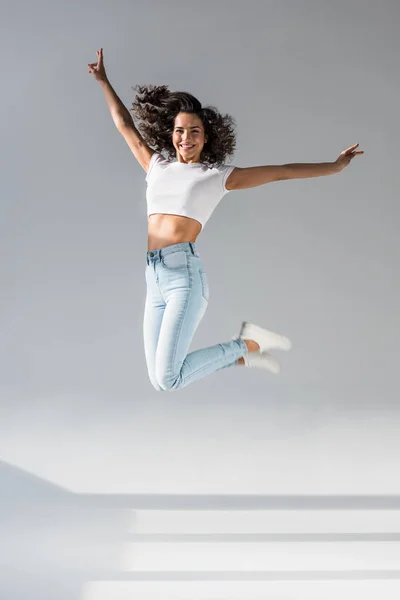 Glückseliges brünettes Mädchen in Jeans lächelt und springt auf grauem Hintergrund — Stockfoto